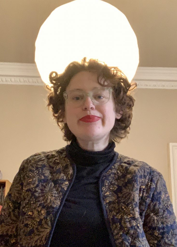 person with red curly hair and red lipstick standing in front of a light that looks like a full moon